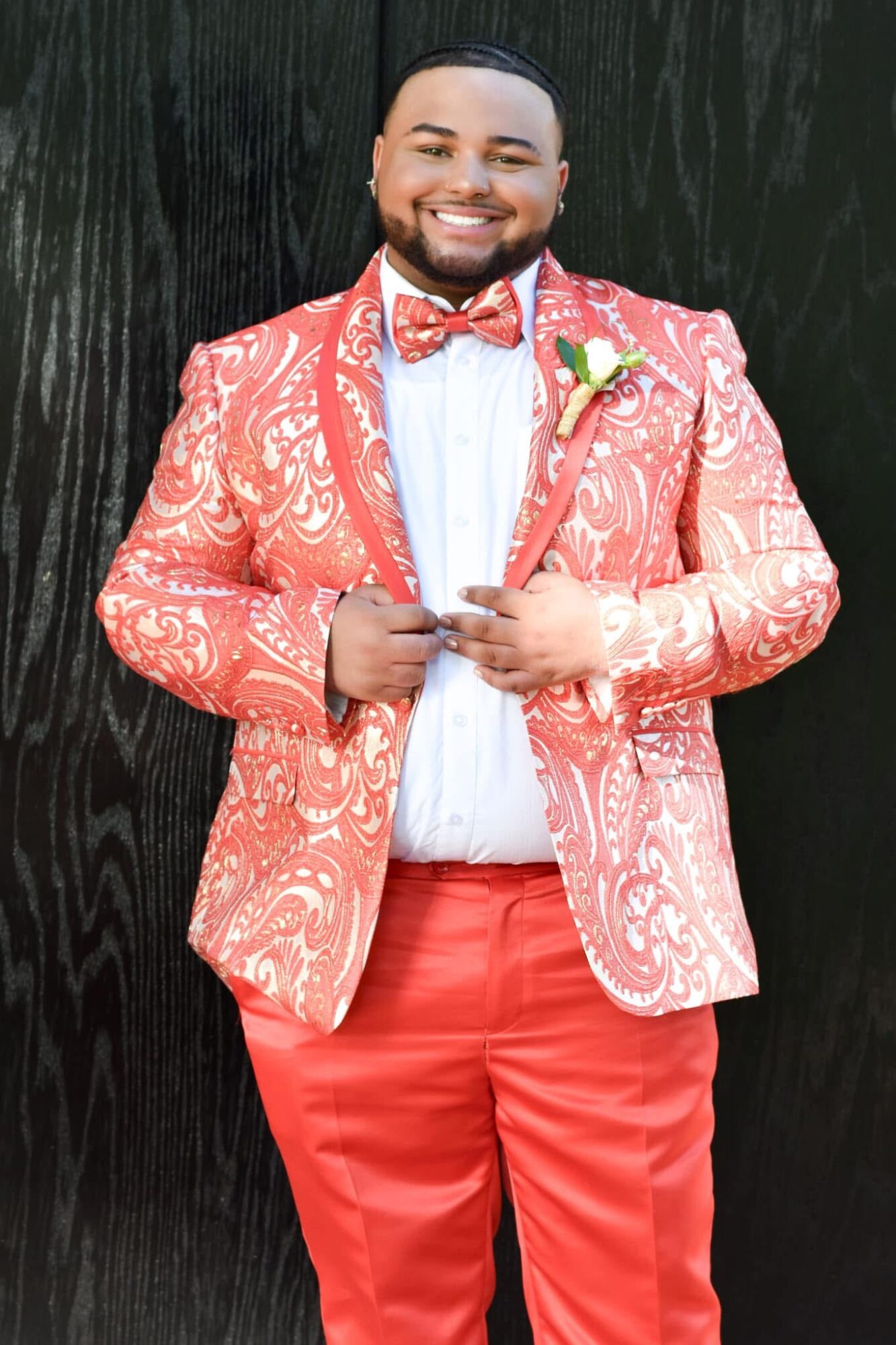 Person smiling, wearing a pink patterned blazer, white shirt, red pants, and a bow tie, standing against a dark background.