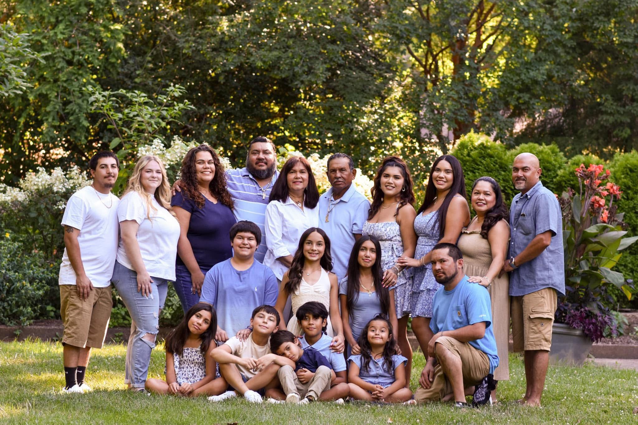 Group of people outdoors in a park, standing and sitting on grass, with trees and flowers in background.