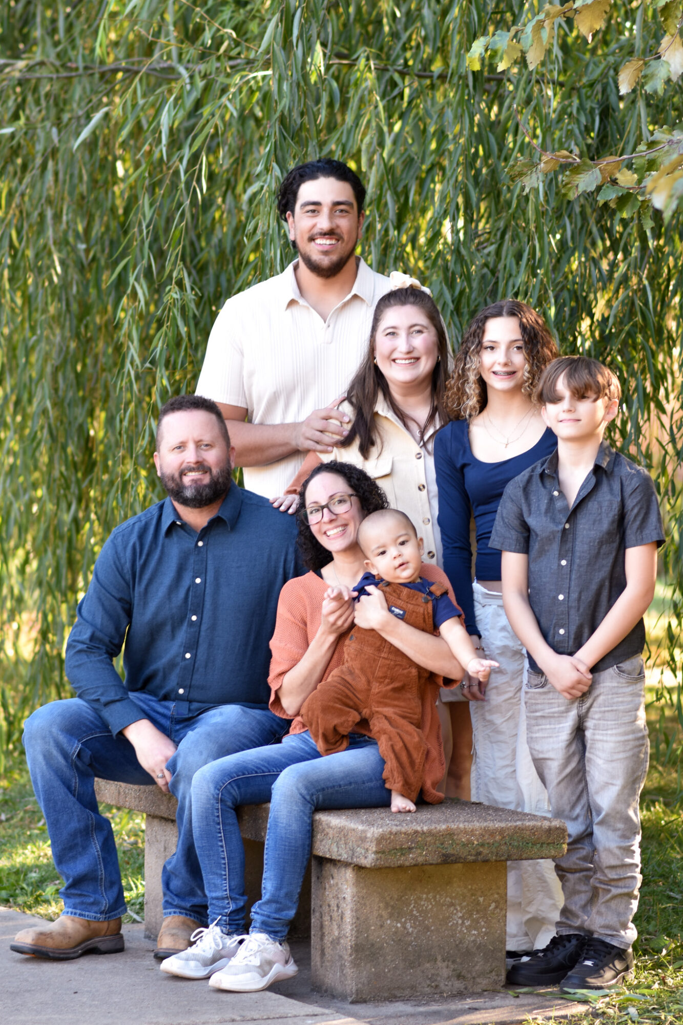 Group of seven people outdoors, standing and sitting on a stone bench, surrounded by tall green plants, smiling.