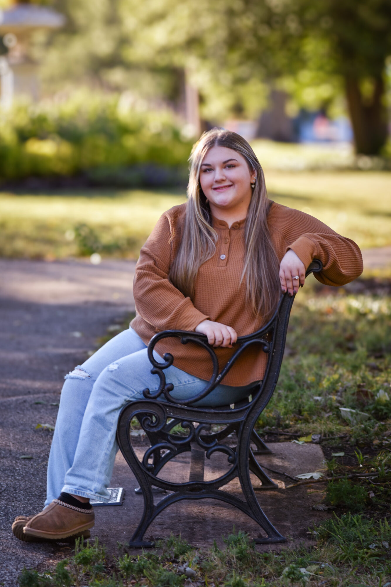 Young woman with long hair sitting on a park bench, smiling, wearing a brown hoodie and light blue jeans.
