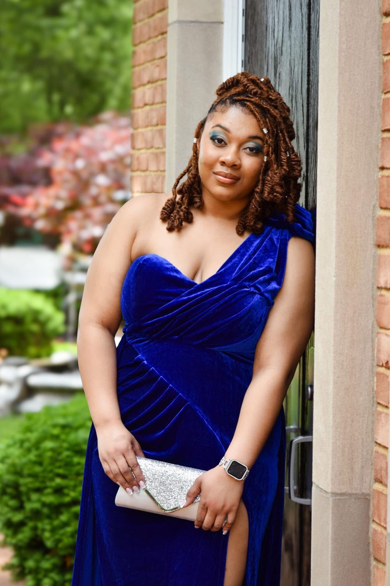 Woman in a blue dress holding a clutch, standing outdoors near a brick wall, smiling.