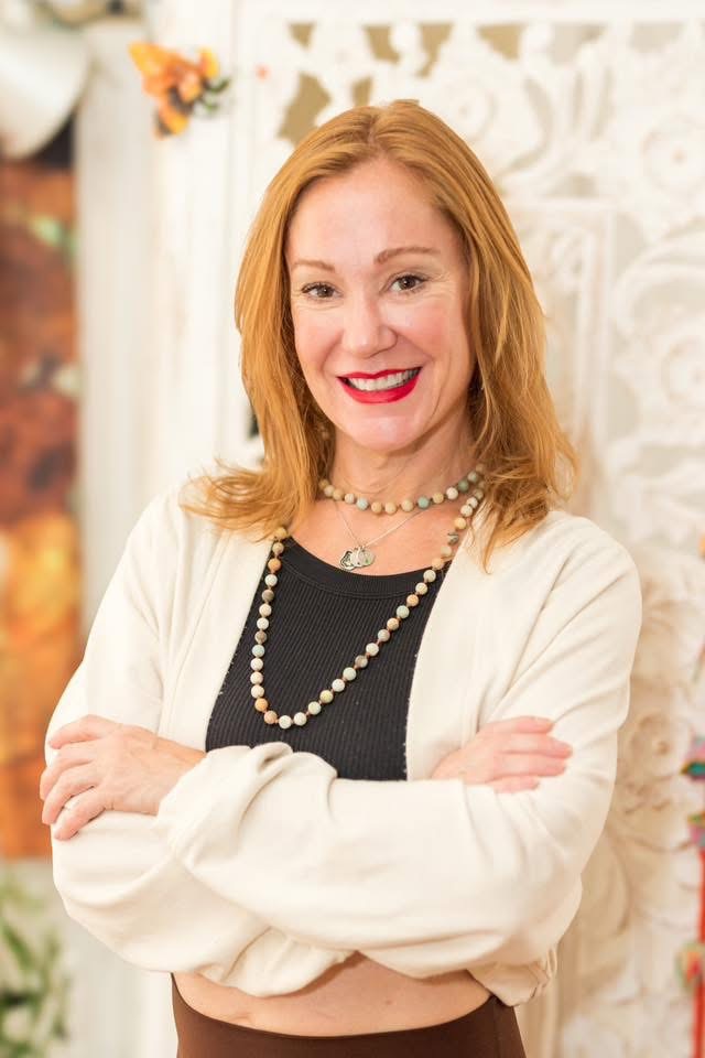 Woman with red hair smiling, wearing a black top, white cardigan, and pearl necklaces, standing with arms crossed in front of a decorative background.