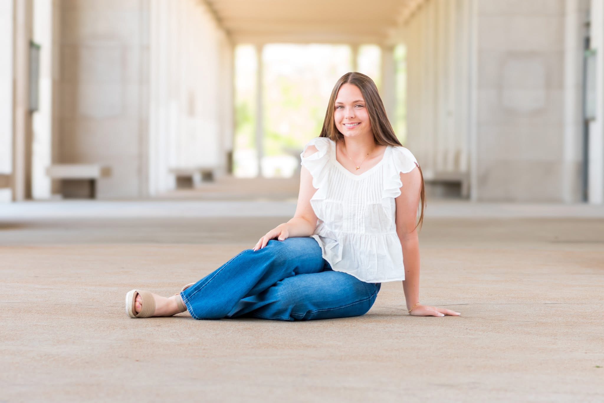 Woman sitting on the floor indoors, smiling, with sunlight streaming through large windows behind her.