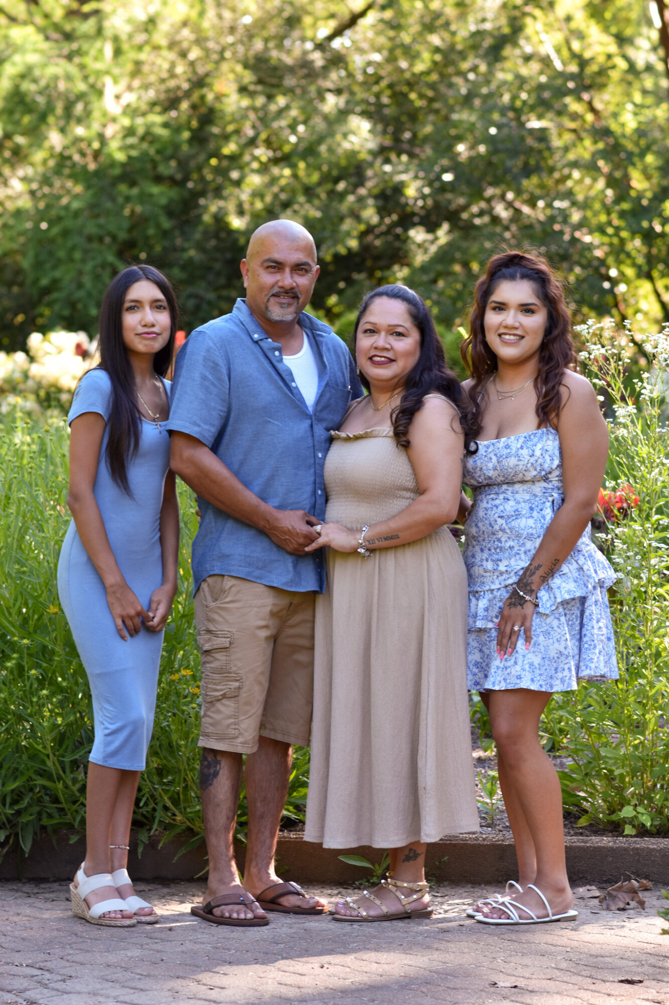 Four people standing outdoors in a park with trees and greenery, smiling, with sunlight filtering through leaves.