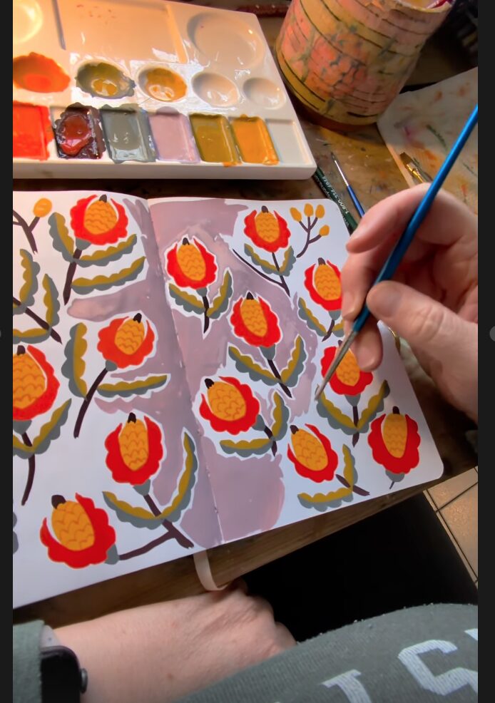 Person painting red and yellow flowers on paper with a small brush, watercolor palette nearby.