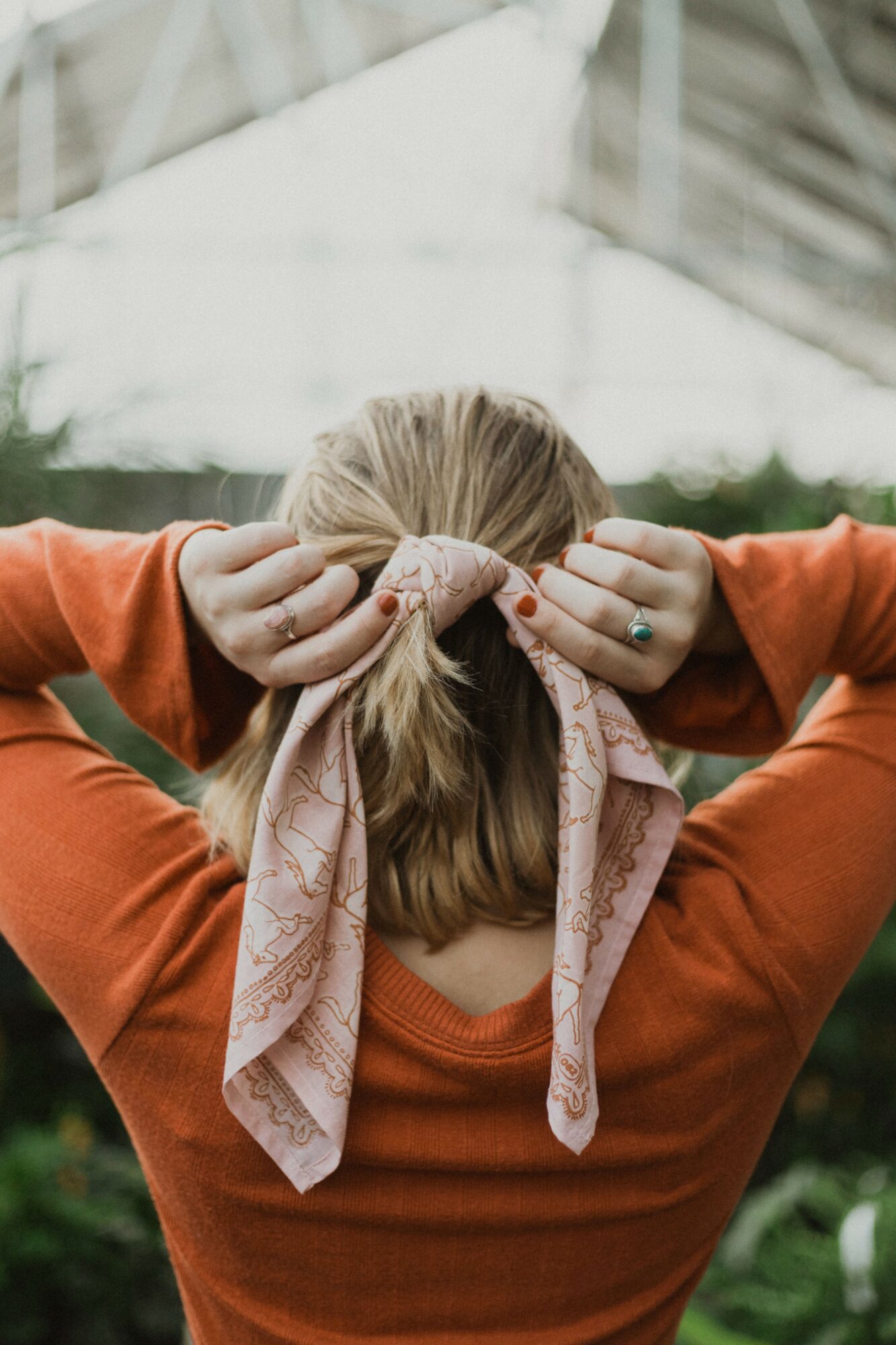 Person with blonde hair tying a pink scarf around their head, wearing an orange top, outdoors with trees and a structure in the background.