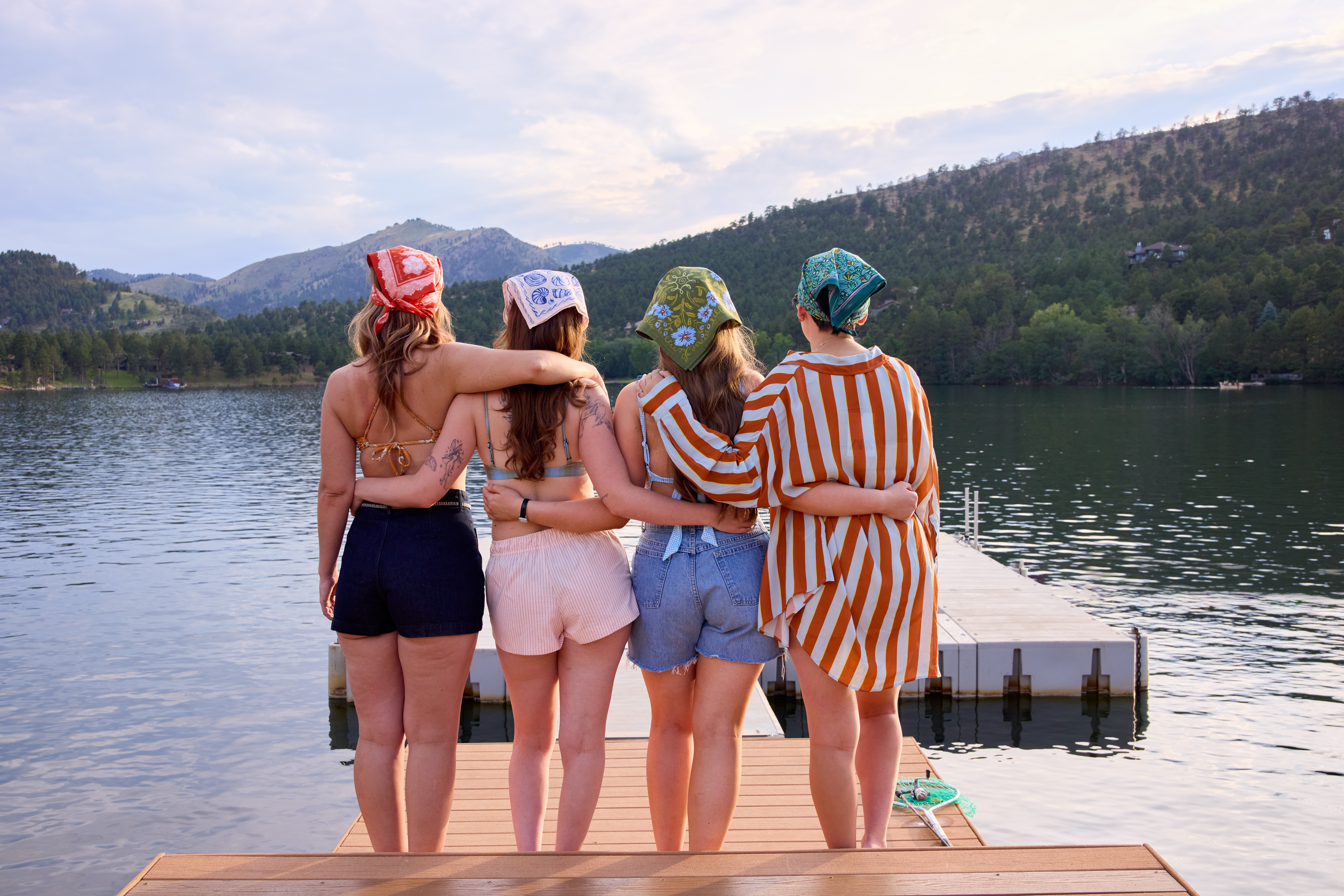 Four girls stand on a dock by a lake, facing away, with mountains and trees in the background.