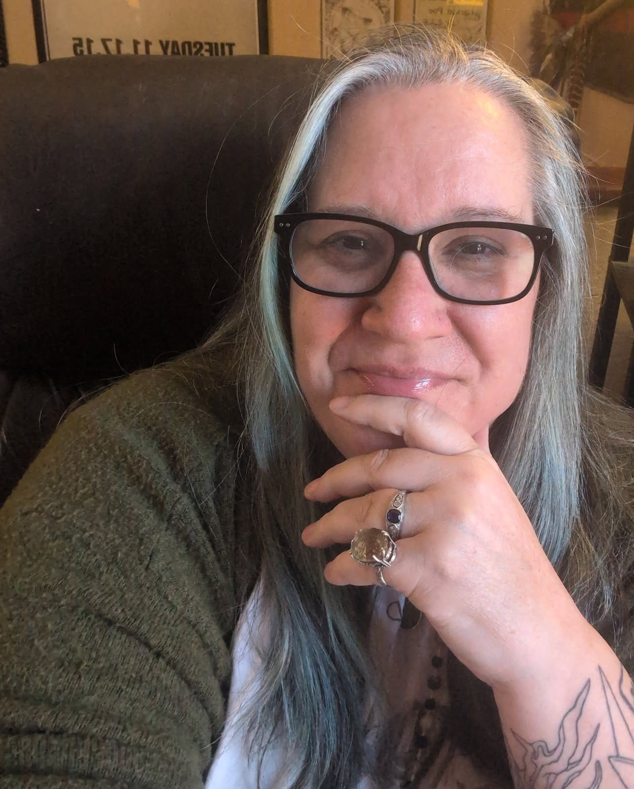 Woman with glasses and long gray hair, resting her chin on her hand, smiling, in an indoor setting.