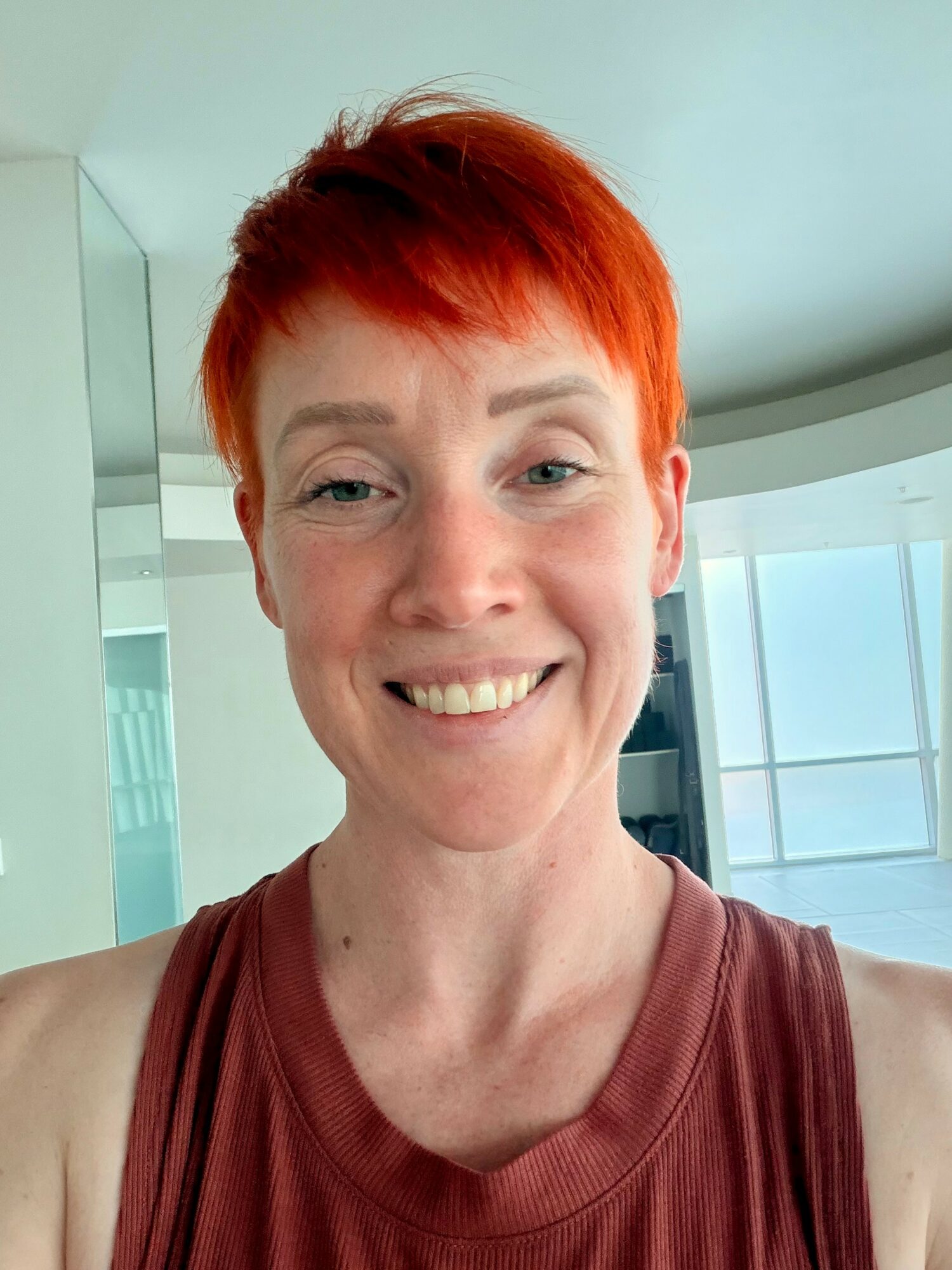 Woman with short red hair smiling, wearing a sleeveless maroon top, in a bright room with large windows.