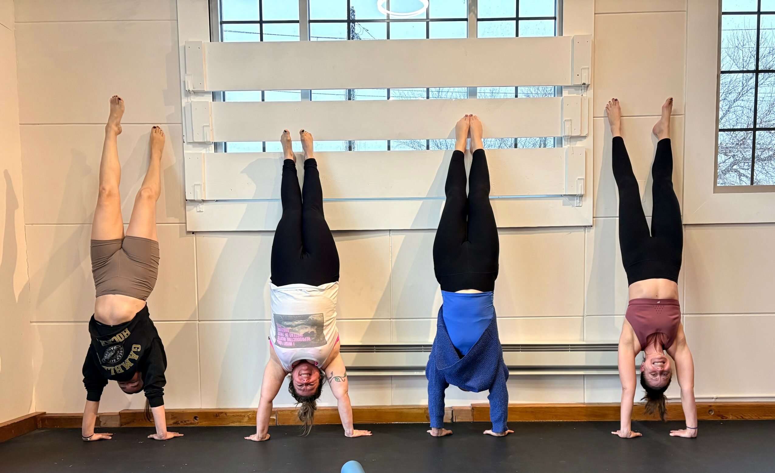 Four people perform handstands against a wall in a room with wooden floors and windows, wearing casual clothing.