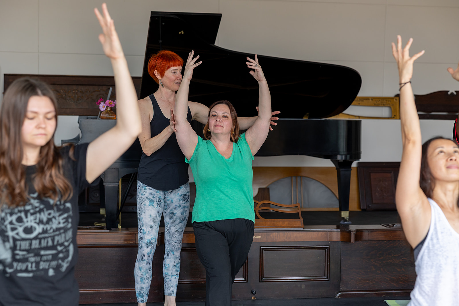 Four women with arms raised, standing on a stage with a grand piano in the background.