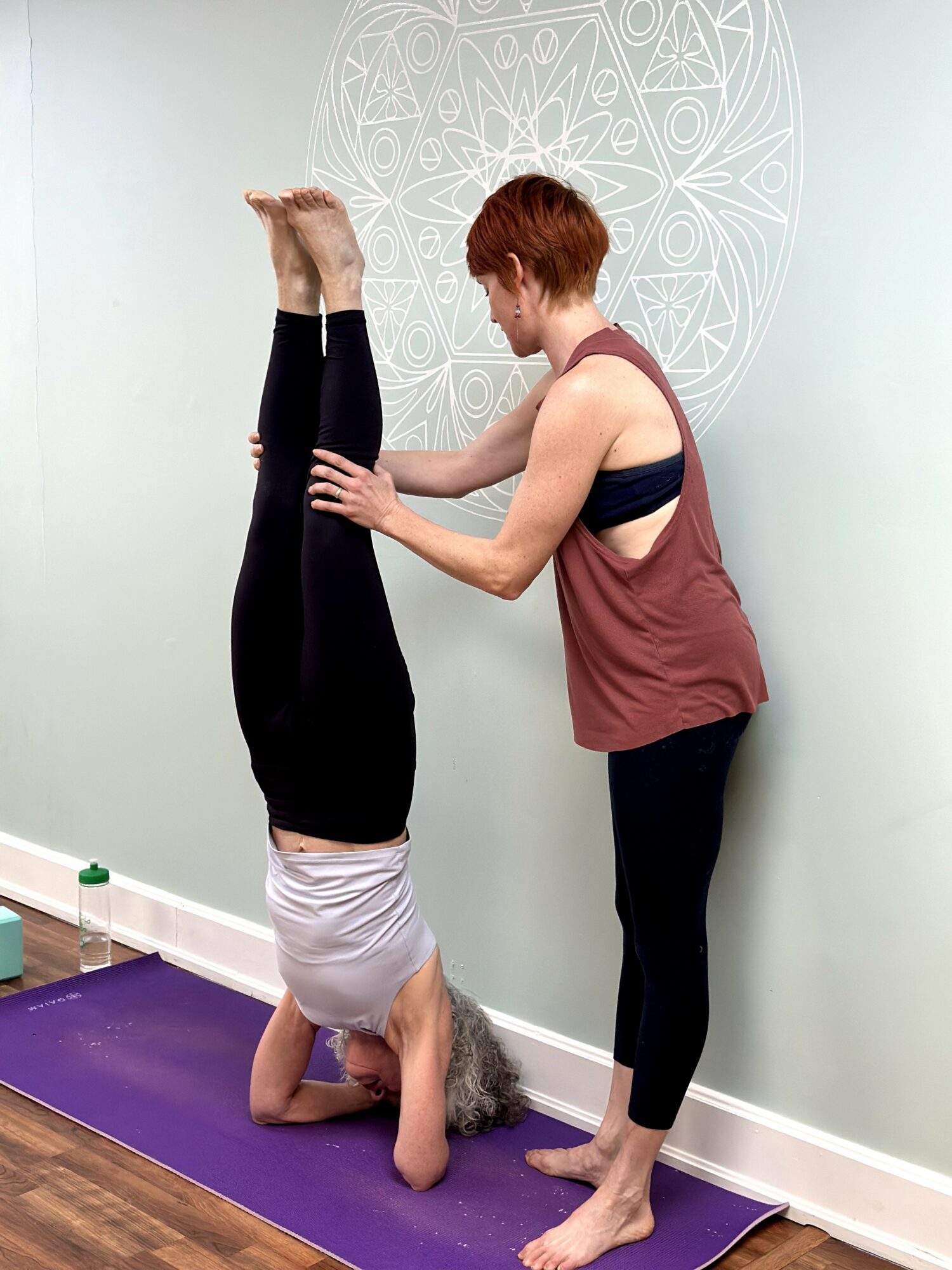 Person performing headstand on yoga mat with instructor assisting, in a room with light green wall and decorative wall art.