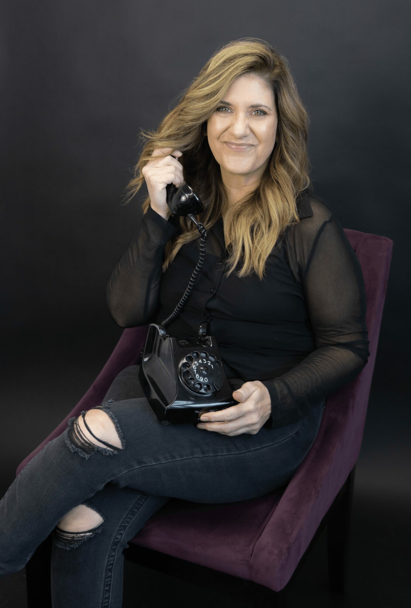 Woman with wavy blonde hair sitting on a purple chair, holding a vintage rotary phone, wearing a black top and ripped jeans.