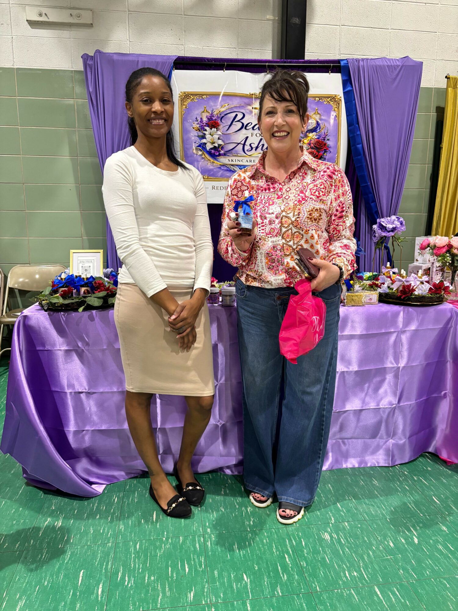 Two women standing in front of a decorated table with purple and pink fabric, flowers, and a sign, indoors.