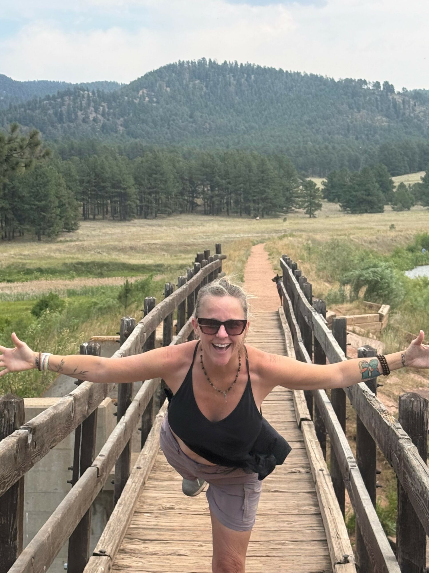 Woman walking on a wooden bridge in a natural landscape with trees and hills in the background.
