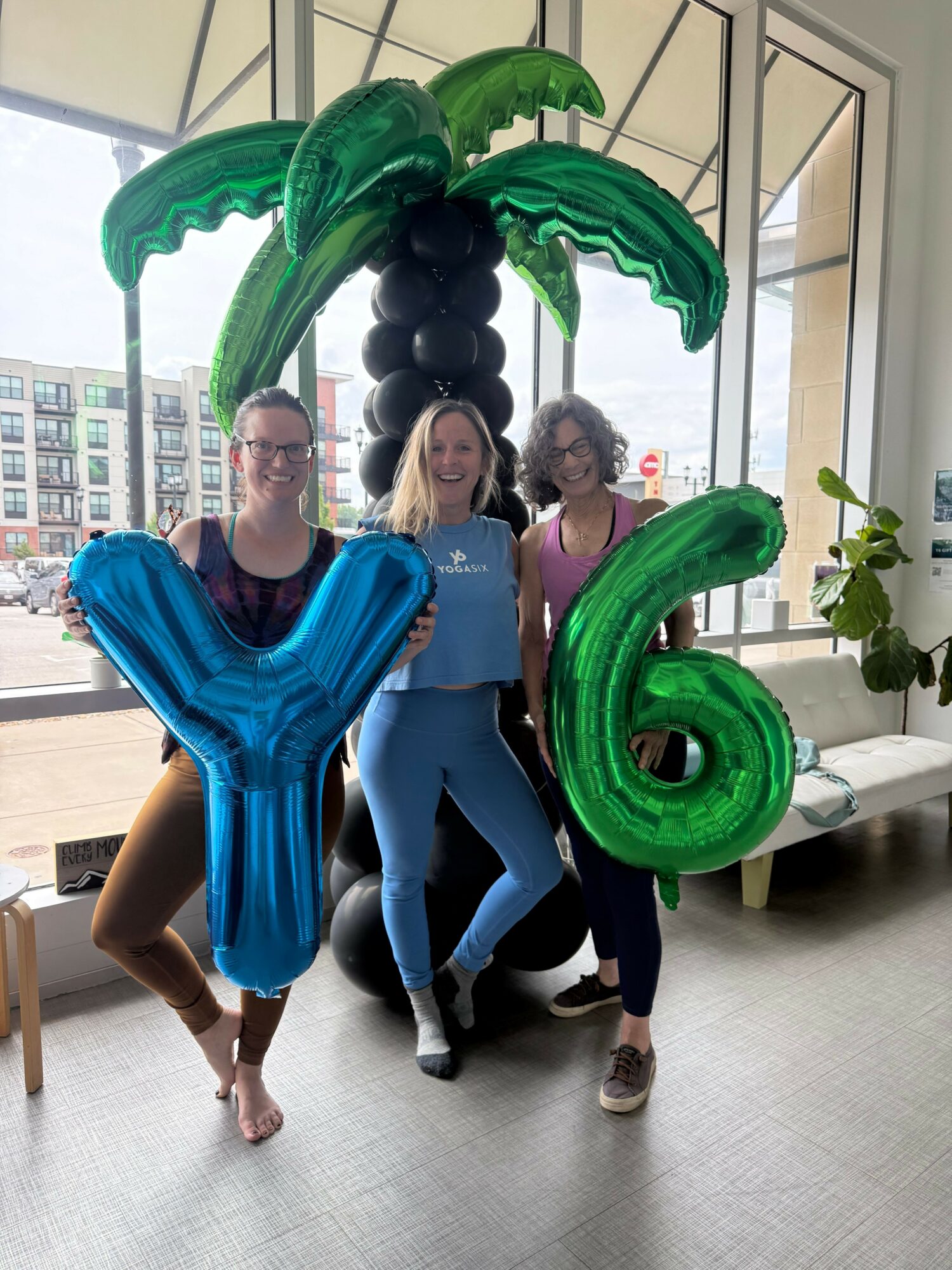 Two women holding large green and blue balloons, standing indoors near a window with a city view, smiling.