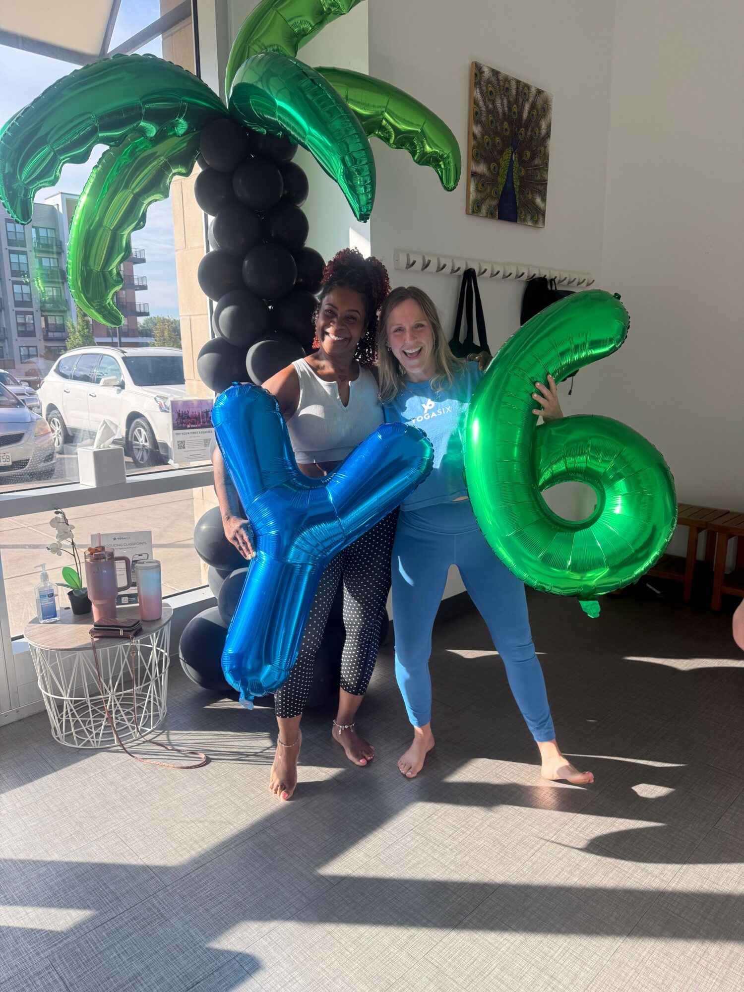 Two people holding large green and blue balloon numbers, with a background of a room and sunlight casting shadows.