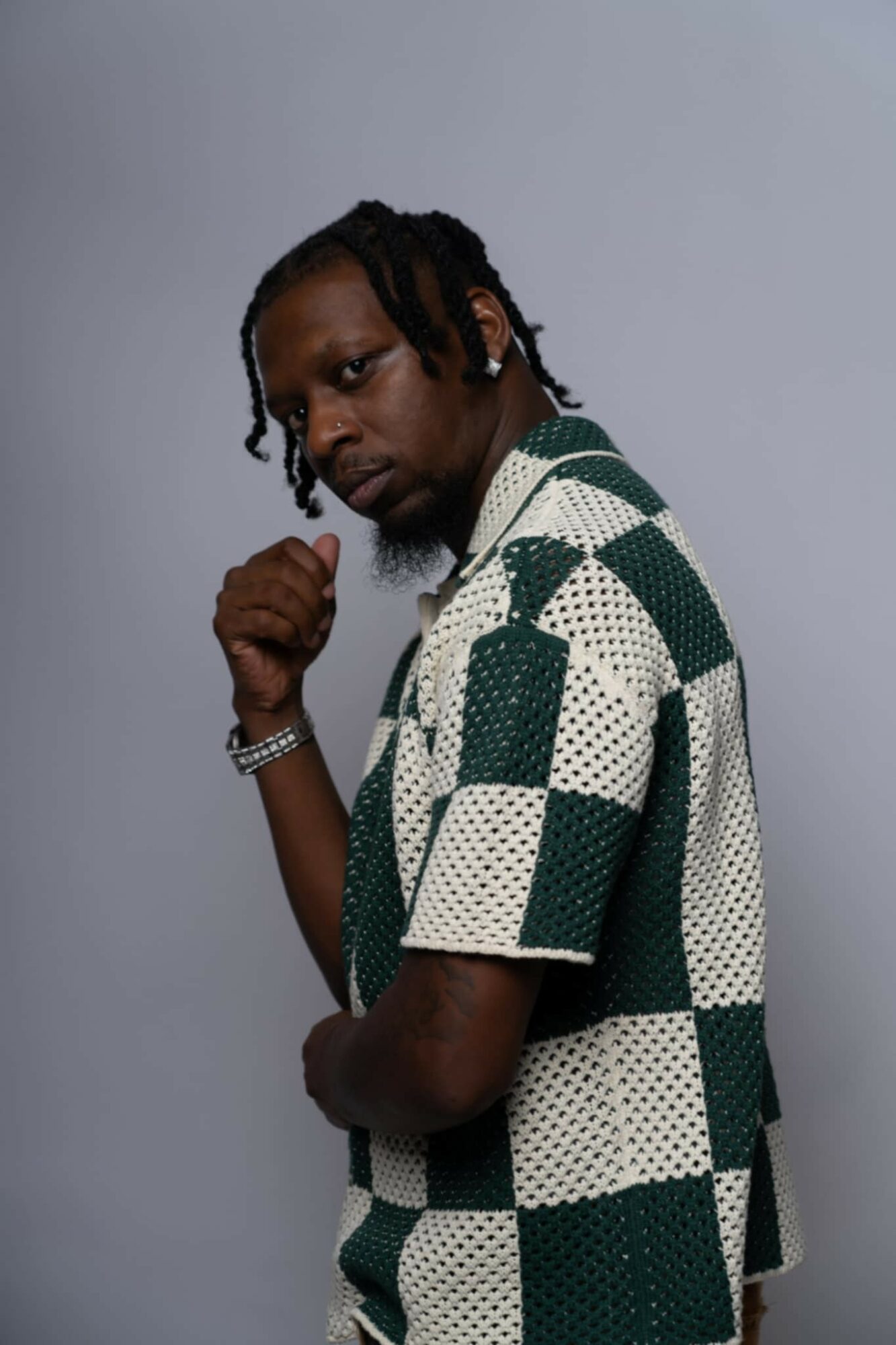 Young man with dreadlocks wearing a checkered shirt, looking at the camera with a serious expression, against a plain background.