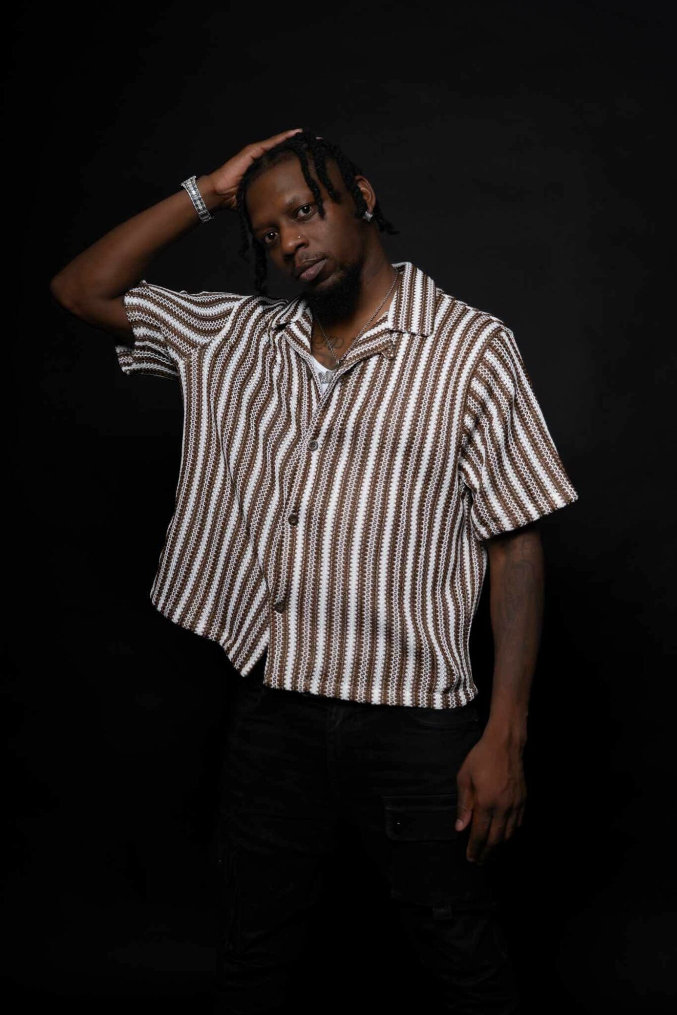 Person with striped shirt and braided hair poses with hand on head against black background.