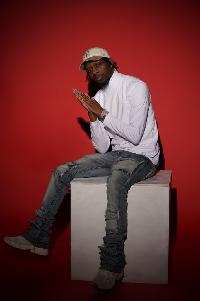 Person sitting on a white block, wearing a white shirt, gray jeans, a white hat, and glasses, against a red background.