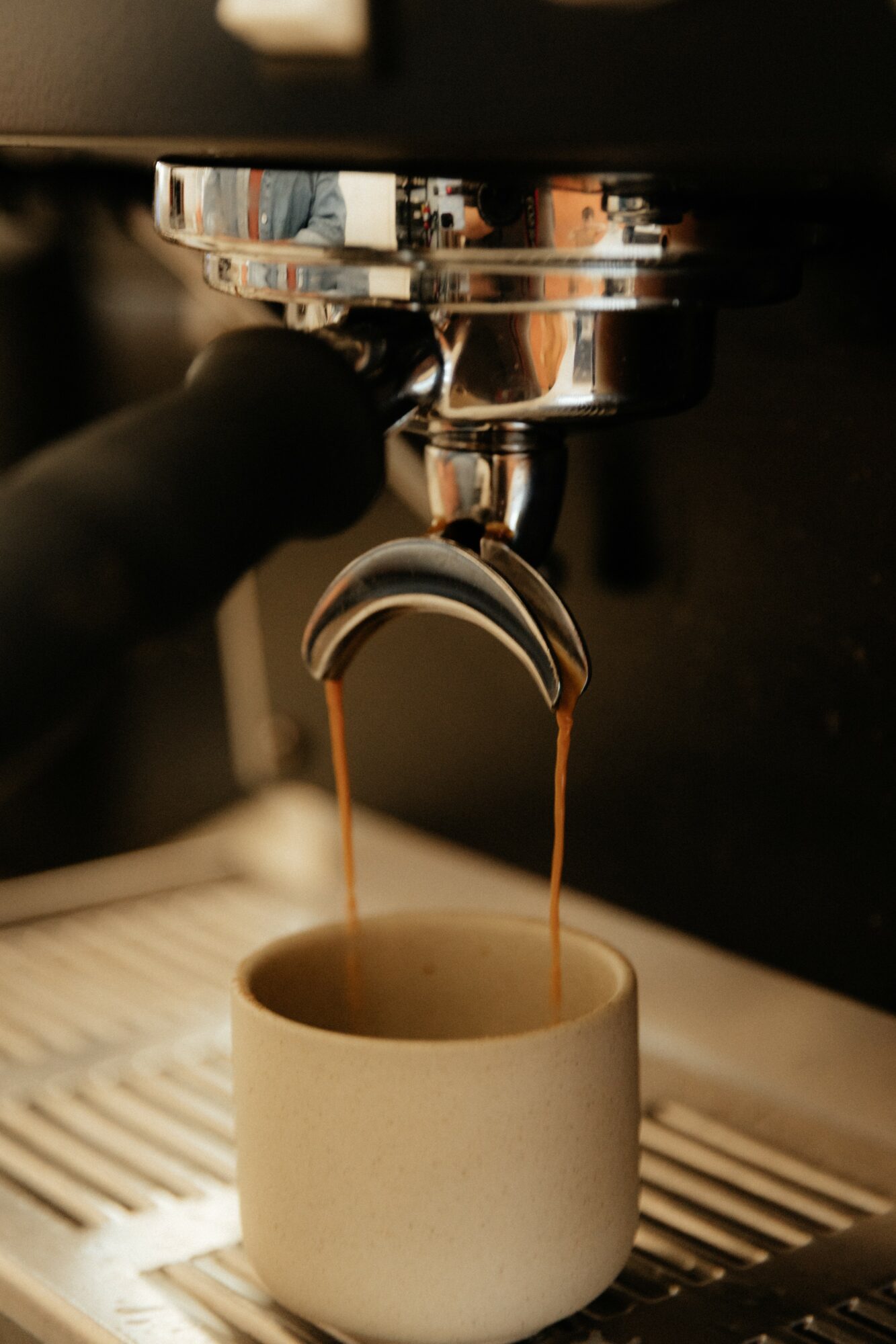 Coffee flows from espresso machine into a white mug on a drip tray, with a hand nearby, in a kitchen setting.