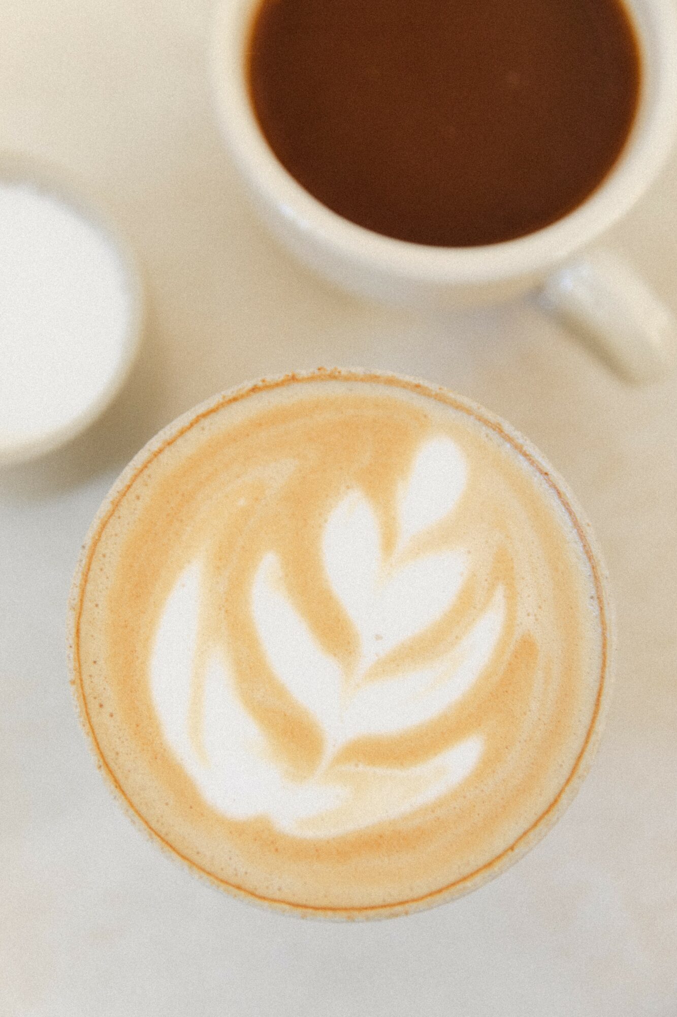 Top view of a latte with foam art, a cup of coffee, and a small container on a light surface.