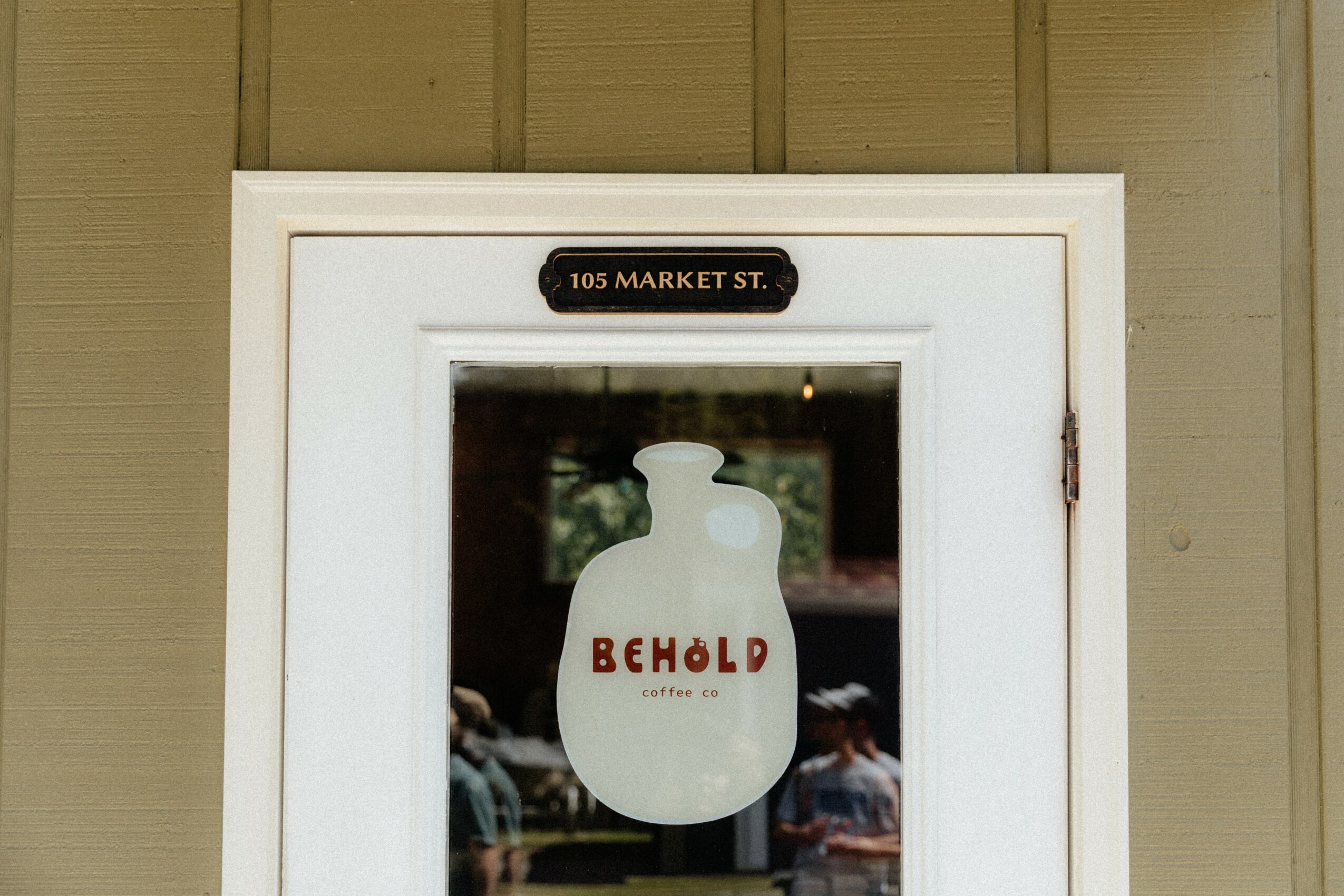 White framed window with a sign for a cafe, featuring a large bottle illustration with the word 'BEHOLD' on it, and a small street address sign above.
