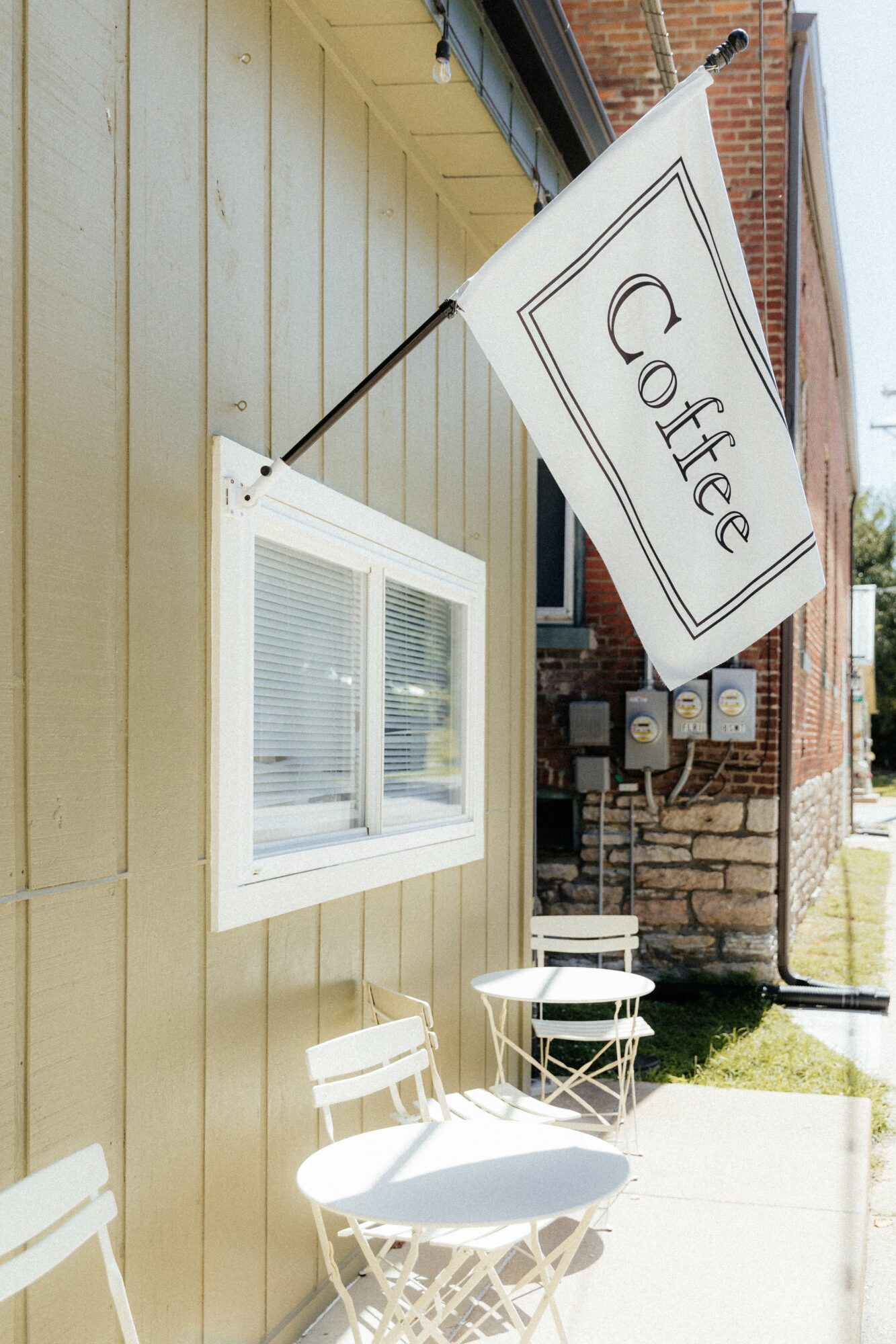 White flag with 'Coffee' written, attached to a building with a window and outdoor chairs, sunny day.