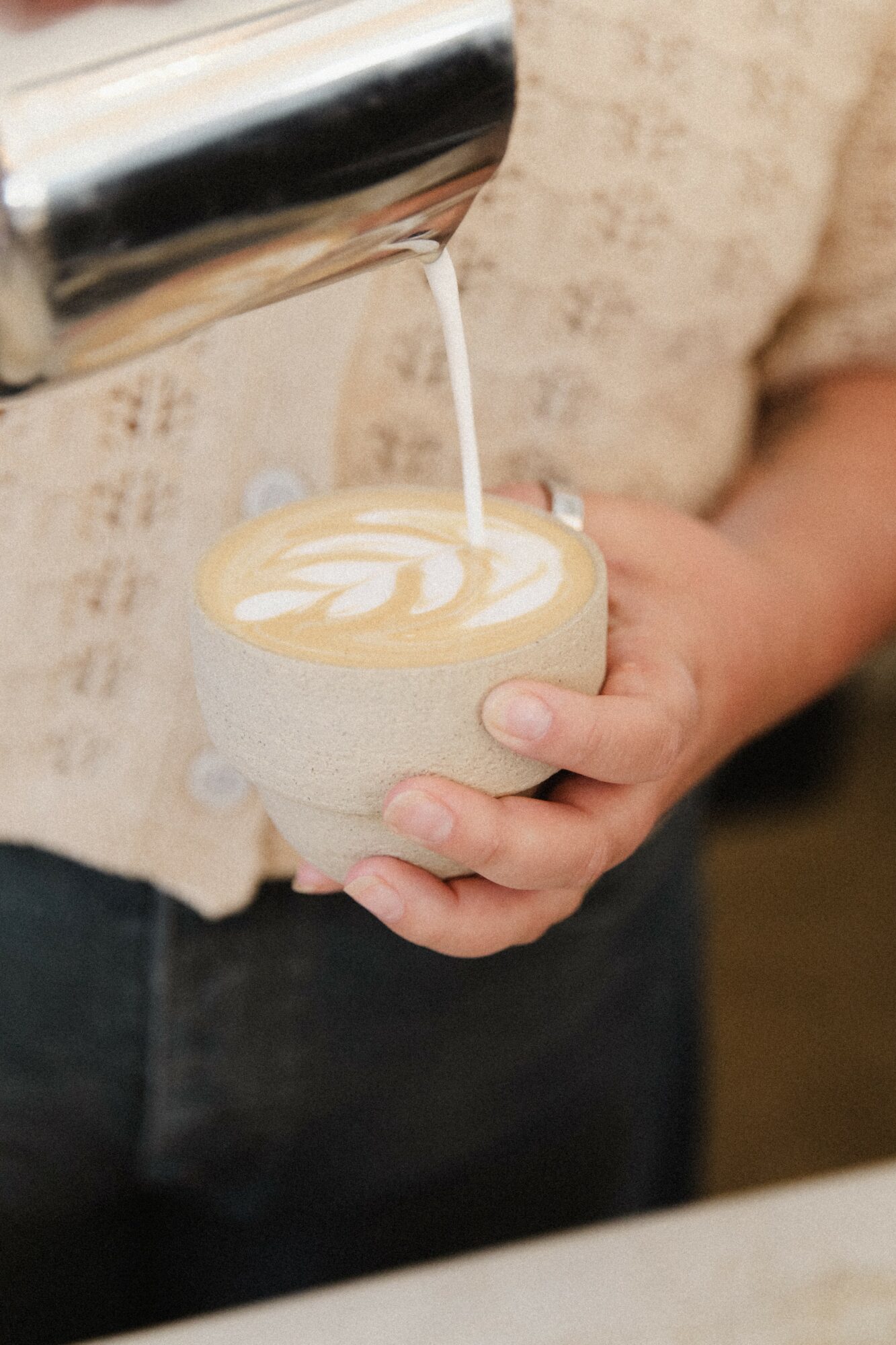 Person holding a cup of latte with latte art, pouring milk from a pitcher, in a cozy setting.