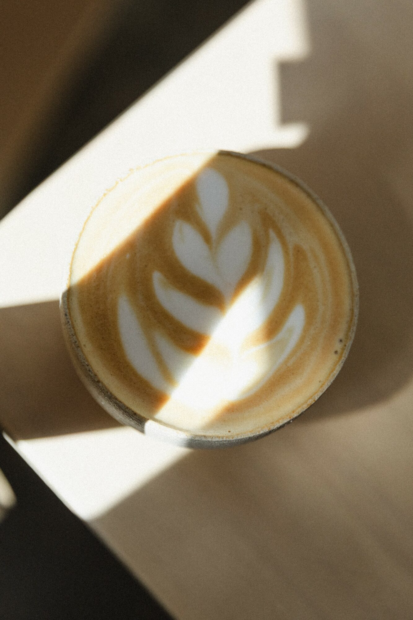 Top view of a latte with latte art on foam, sunlight casting shadows, in a cup on a table.