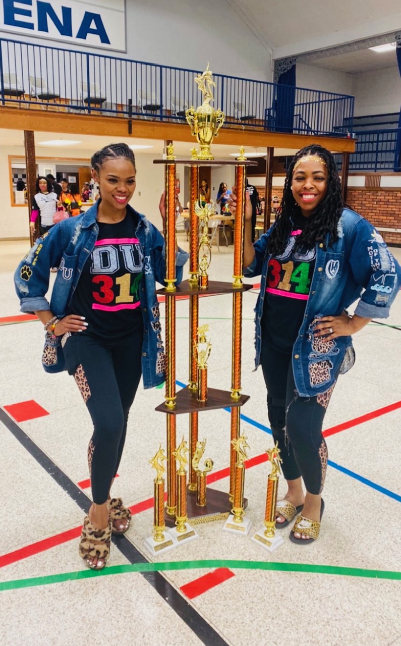 Two women stand on a gymnasium floor holding a large trophy, smiling, with a gymnasium and spectators in the background.