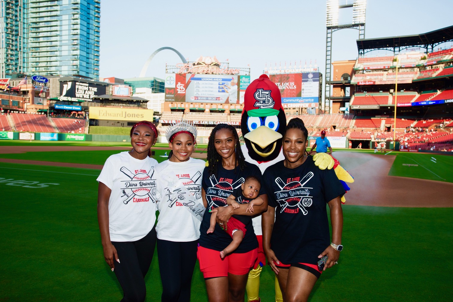 Four women and a mascot pose on a baseball field with a city skyline in the background, smiling and holding a baby.