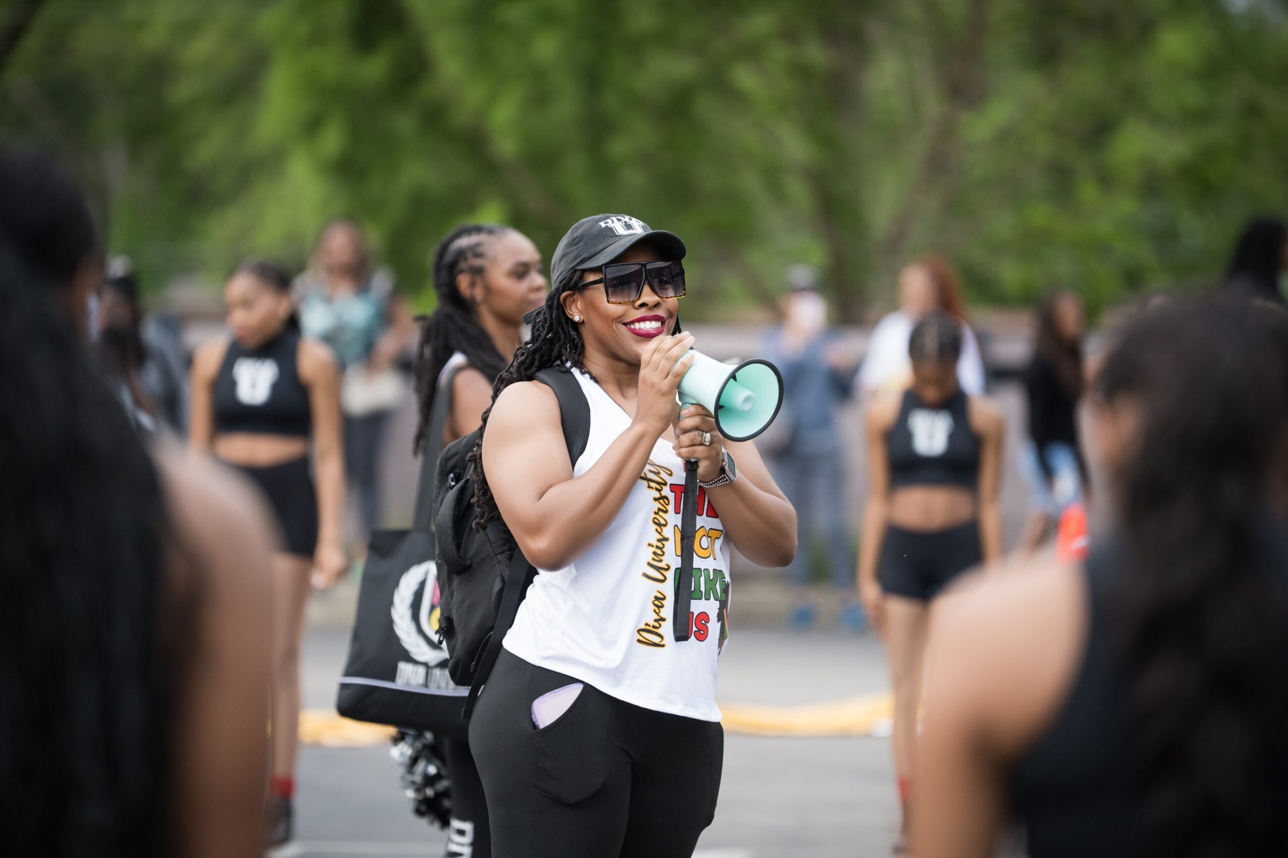 Woman with sunglasses and a megaphone speaking to a group outdoors, with trees in the background.