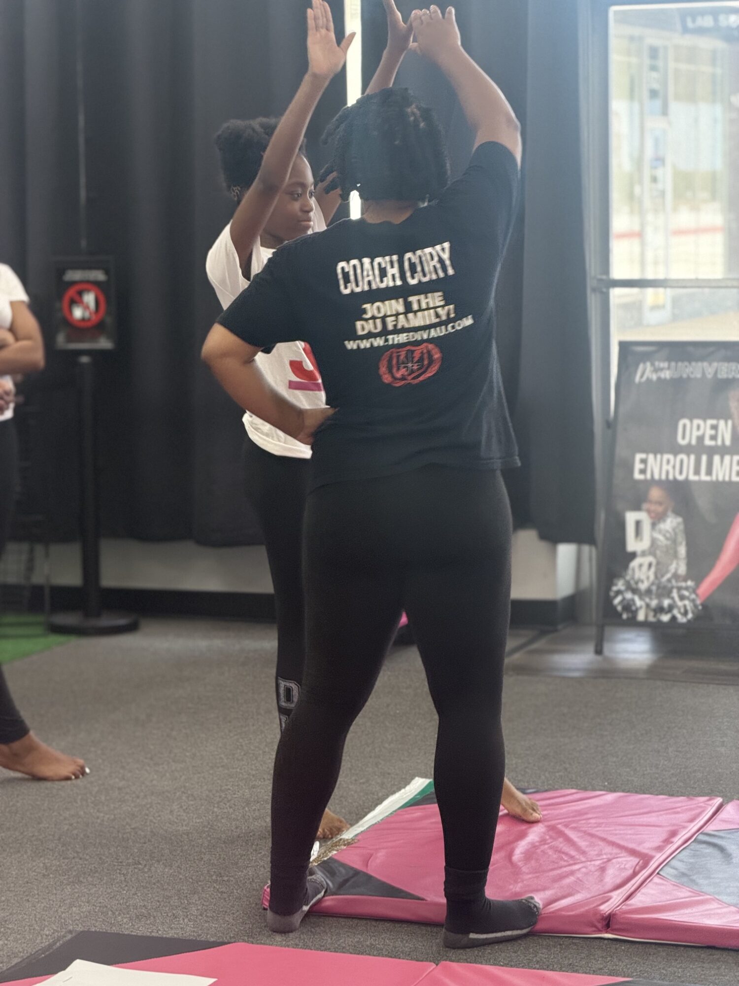Person standing on a pink mat with arms raised, wearing a black shirt and black pants, indoors near a window.