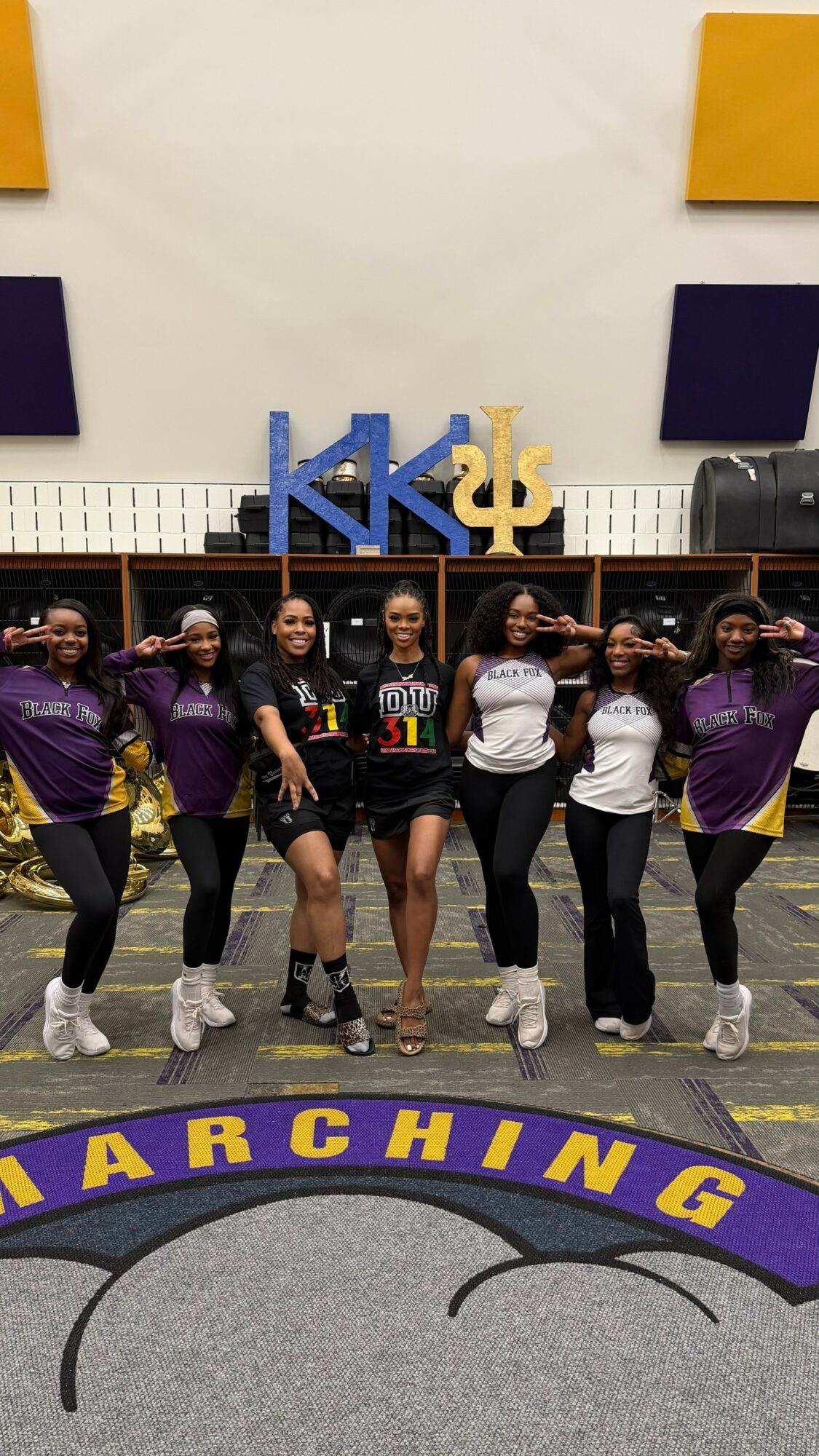 Six women in purple and white shirts stand in a line, saluting, in a gym with Greek letters and a banner.