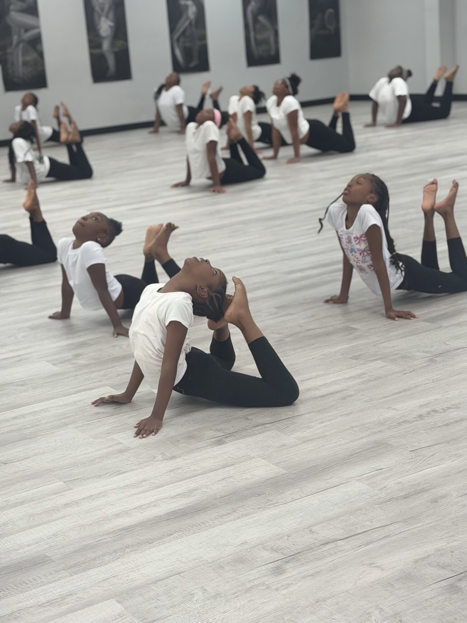 Children in a dance studio practicing poses on the floor, some with legs raised and others supporting with hands, all wearing white tops and black bottoms.