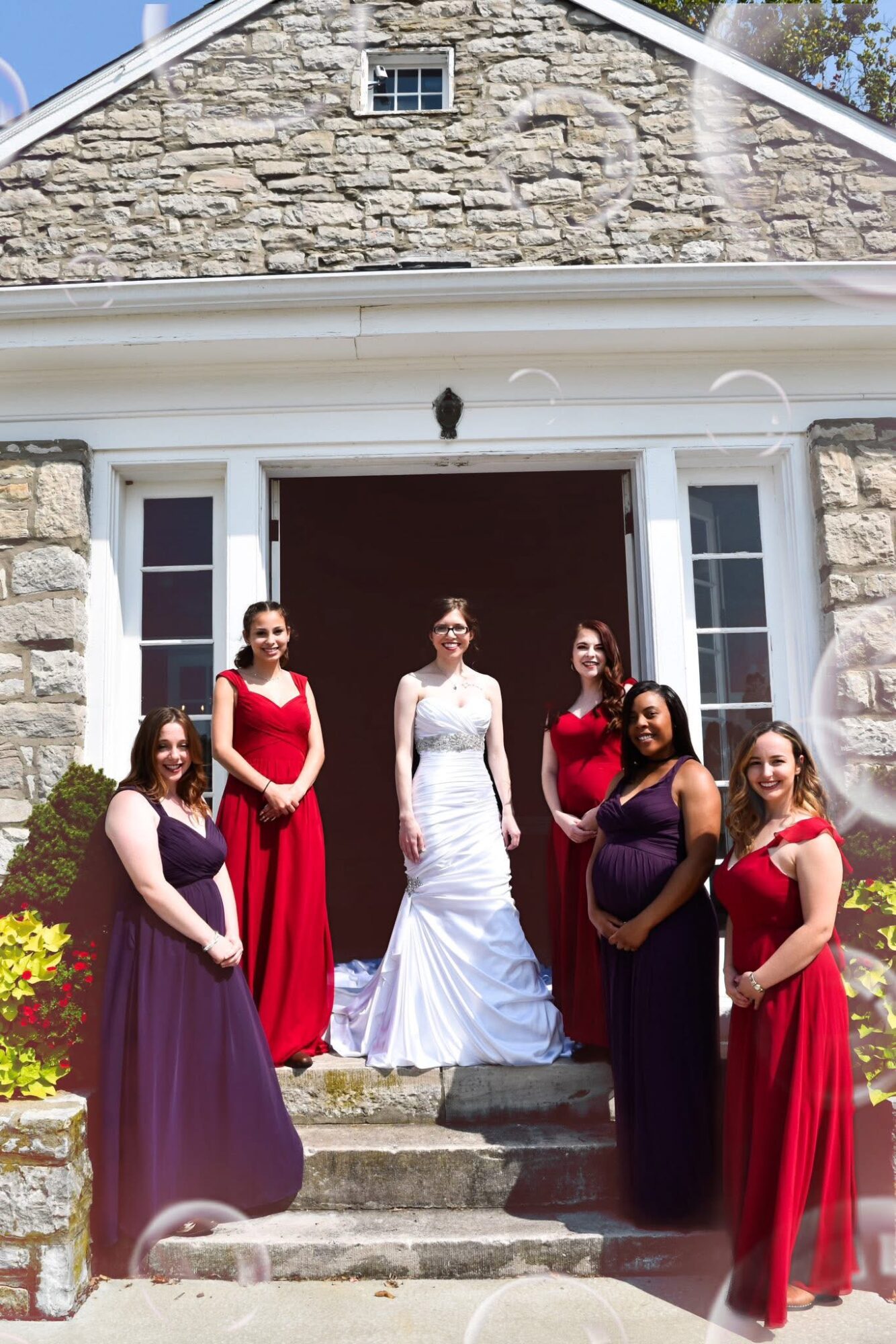 Six women in formal dresses stand on steps in front of a stone building with white trim and a large open door.