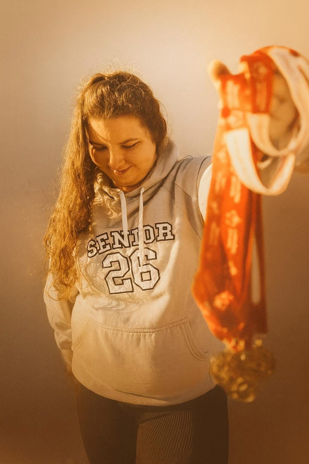 Young girl with long curly hair holding a colorful cloth, looking down, wearing a hoodie with 'Senior 26' on it.