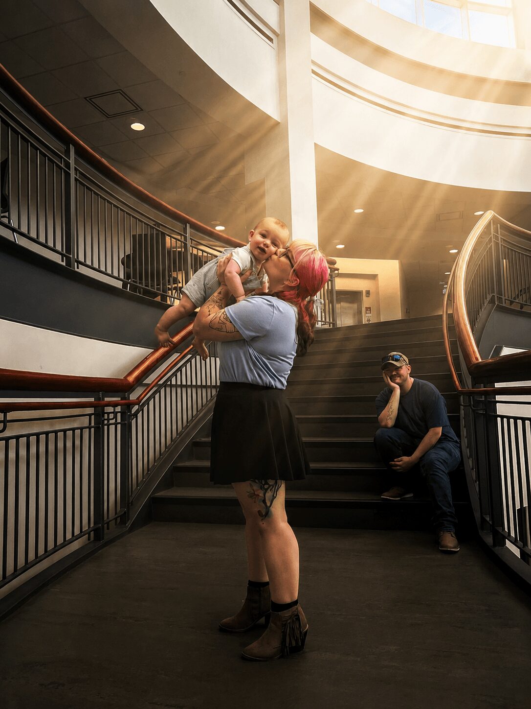 Person lifting another person with pink hair in a circular indoor space, sunlight streaming in from above, person sitting on stairs with camera