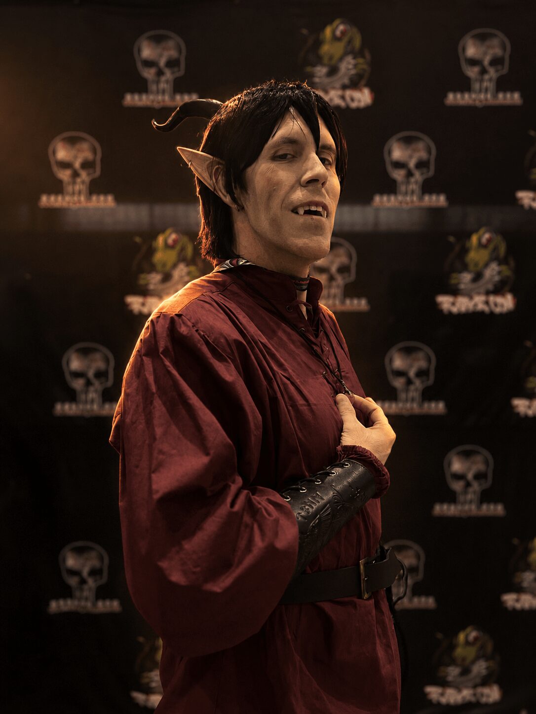 Person dressed as a fantasy character with pointed ears, dark hair, and a maroon outfit, standing in front of a themed backdrop.