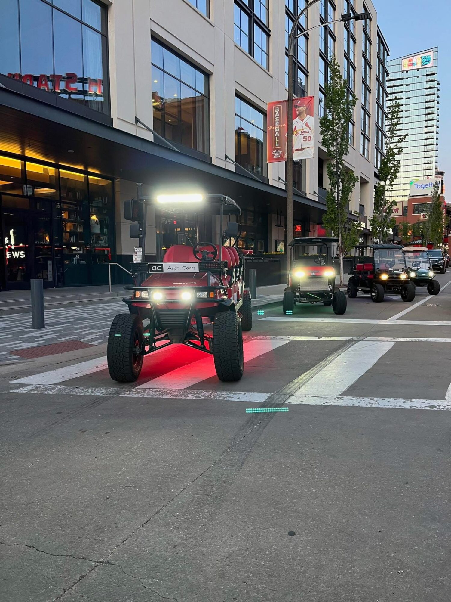 Off-road vehicle with bright headlights parked on city street at dusk, buildings and other vehicles in background.