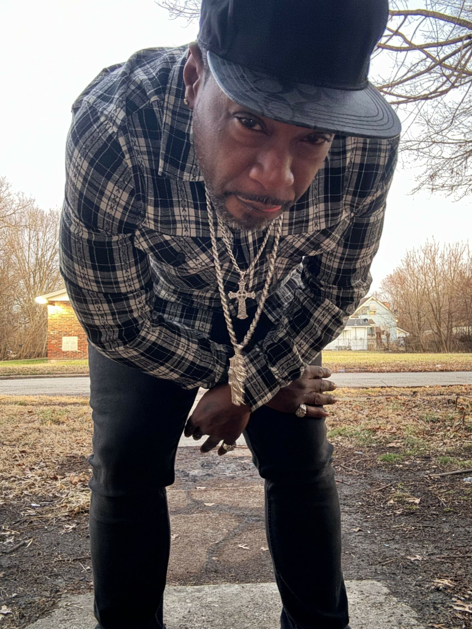 Man wearing a plaid jacket, black hat, and gold chain, standing outdoors on a dirt path with trees in the background.