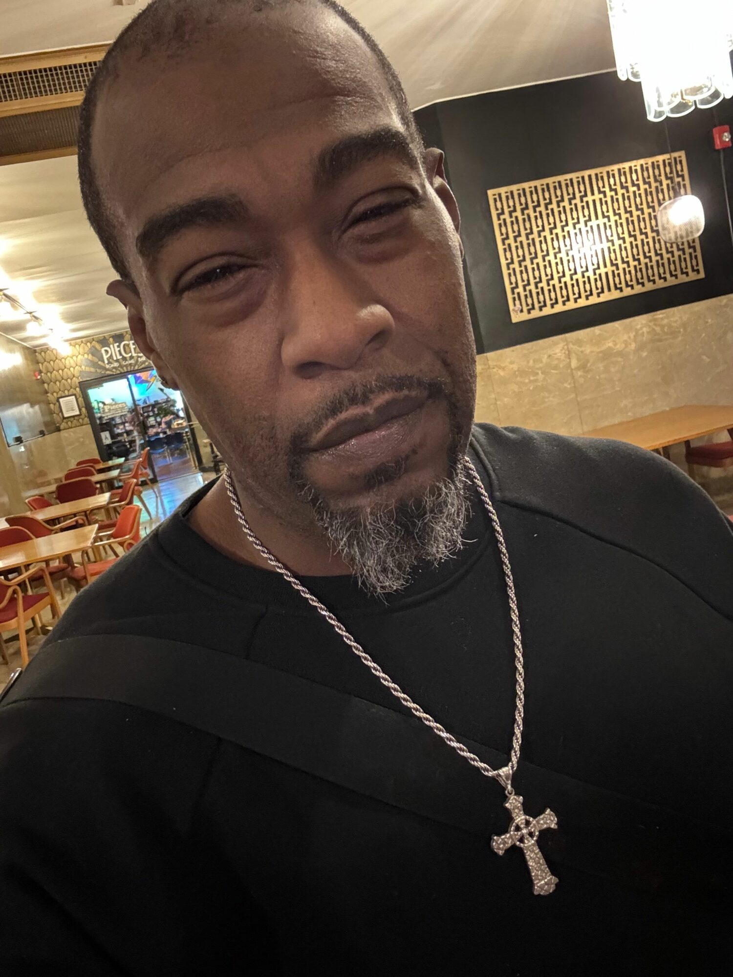 Man with a beard and chain necklace in a restaurant or cafe setting, smiling with eyes closed.