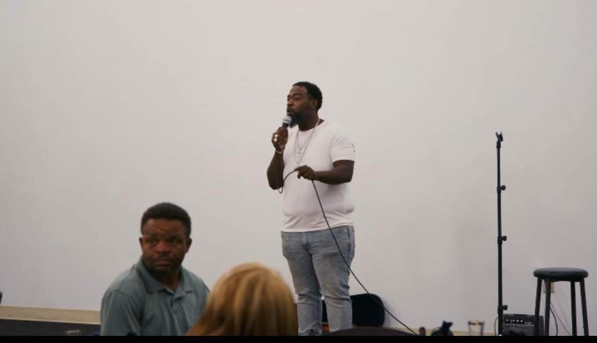 Man standing on stage holding a microphone, with a person sitting nearby and a stool on the right.