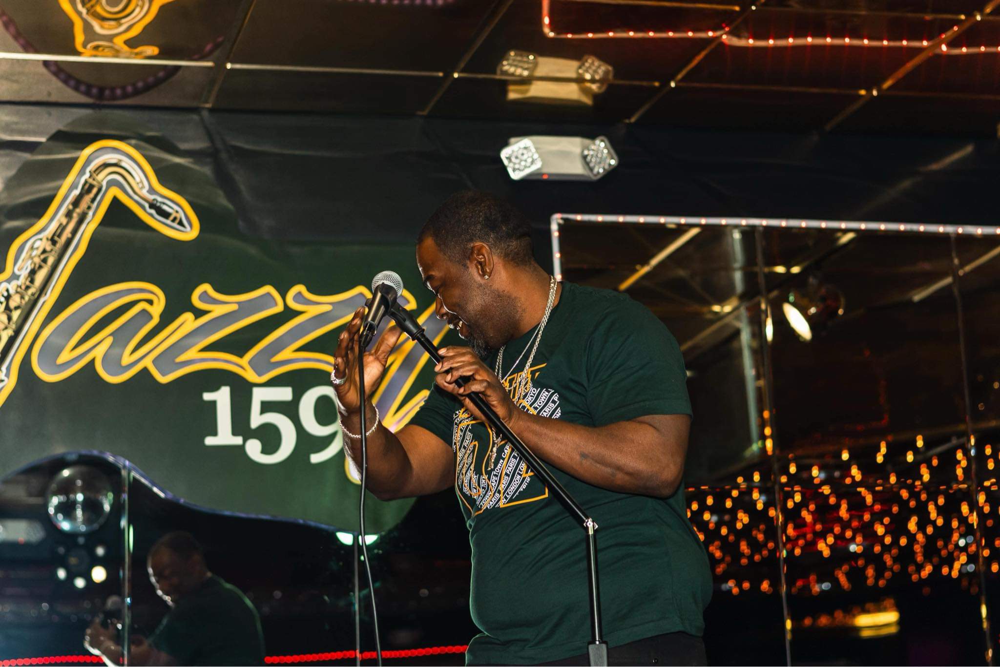 Person singing into a microphone on stage with a dark background and decorative lights, smiling and holding the microphone with both hands.