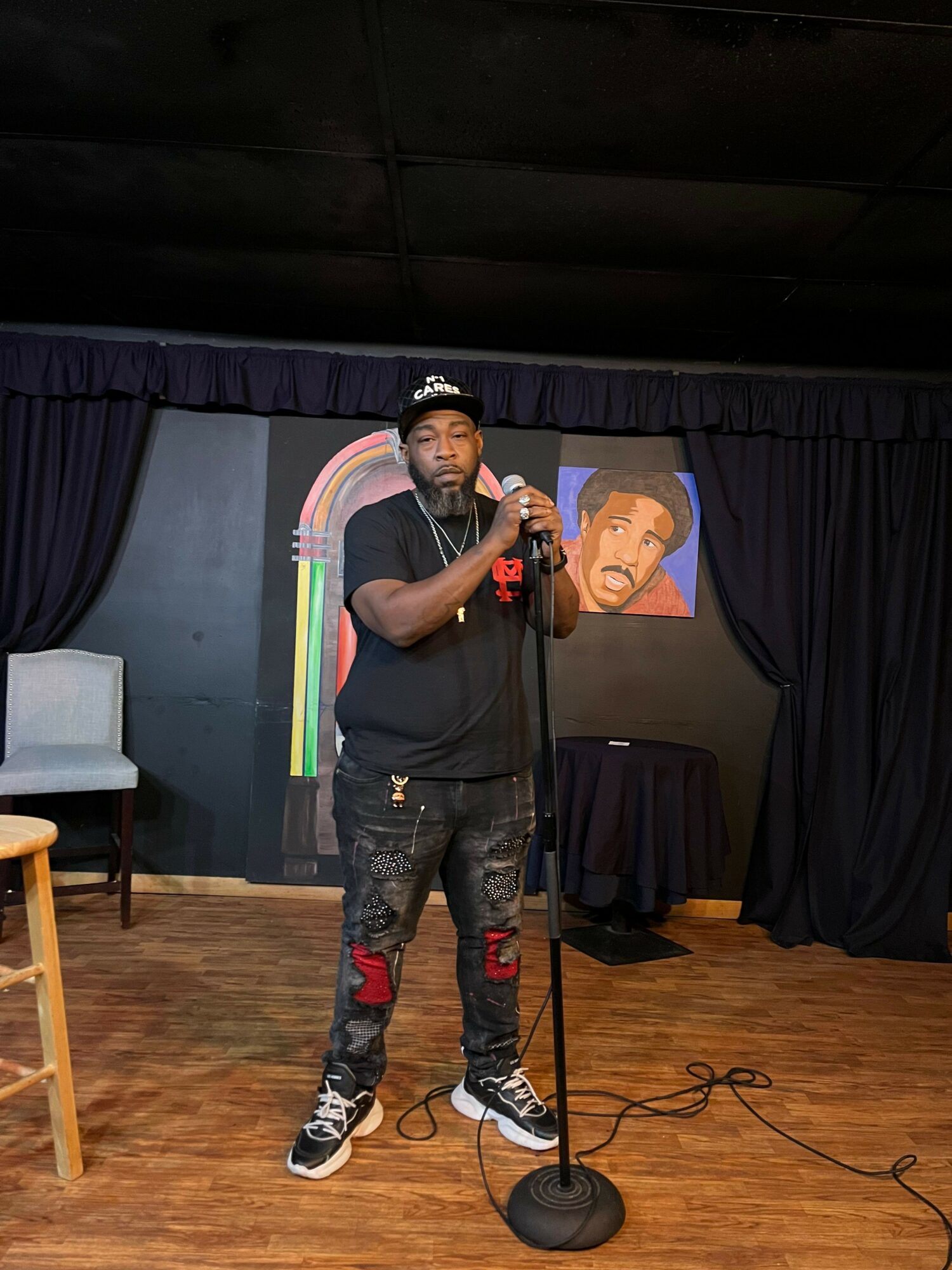 Person standing on stage with microphone, wearing black shirt and jeans, in front of a colorful backdrop and painting, wooden floor.