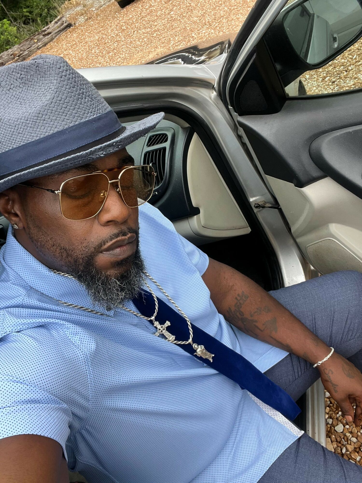 Man with sunglasses and a hat lying inside a vehicle, wearing a light blue shirt, with a strap across his chest.
