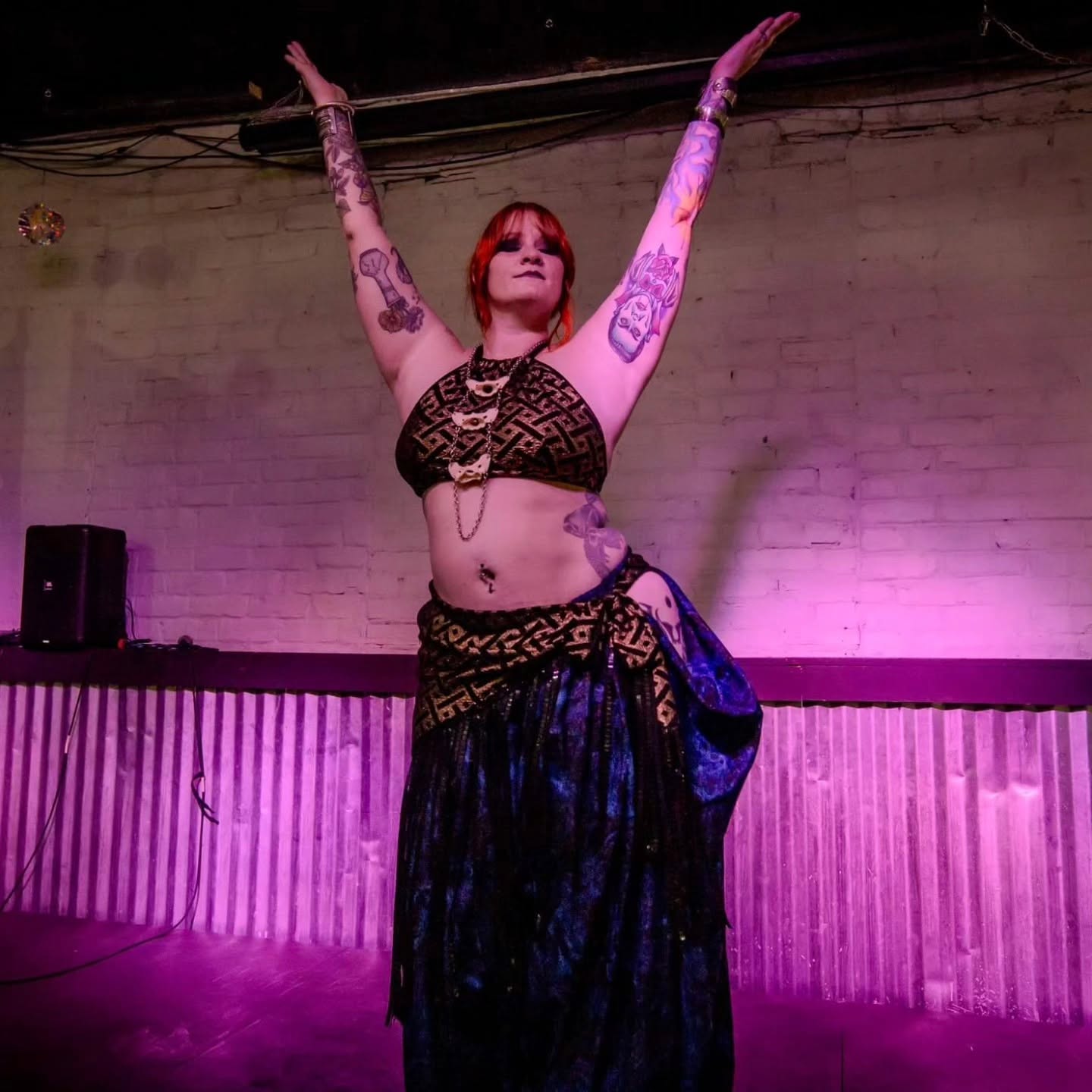 Person with tattoos and red hair raises arms, wearing a black top and long skirt, on stage with purple lighting.