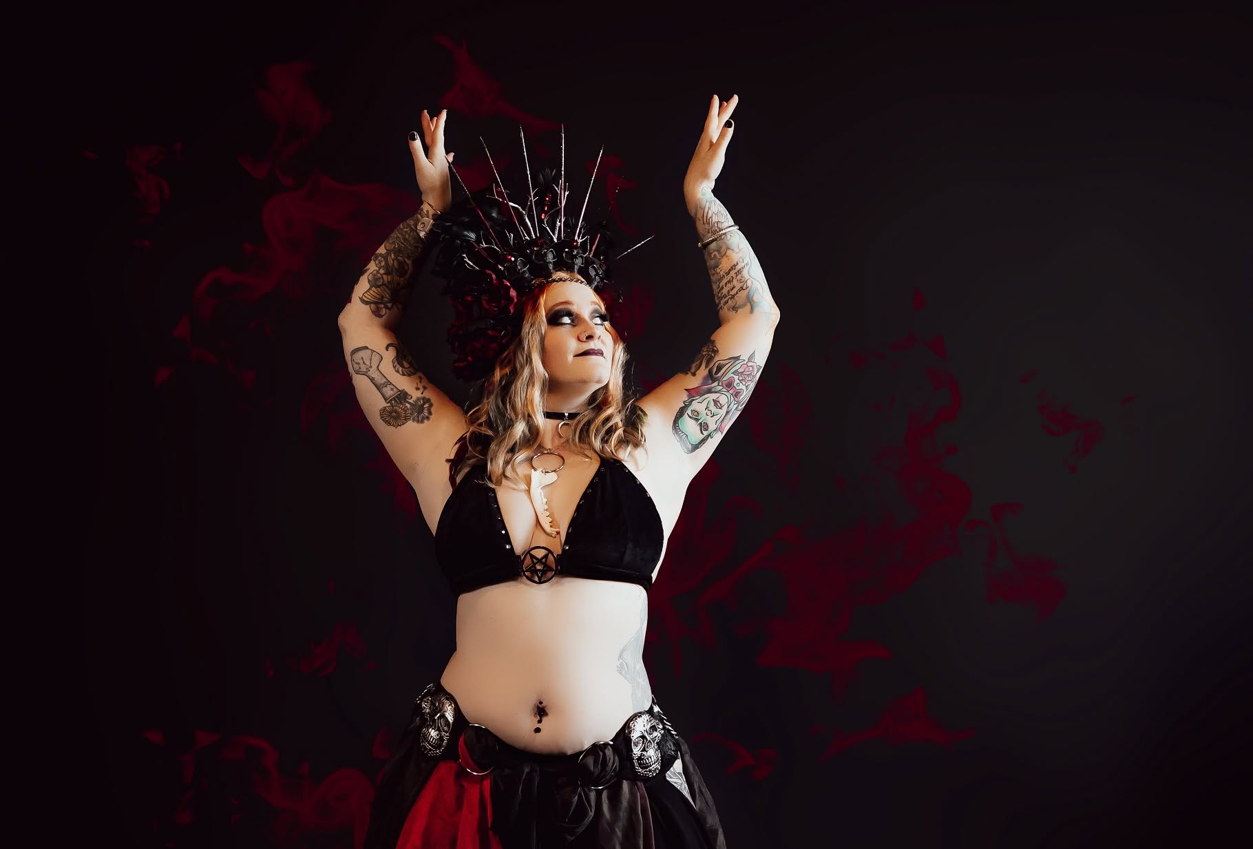 Woman with tattoos wearing a crown and black and red outfit, raising her arms against a dark background.