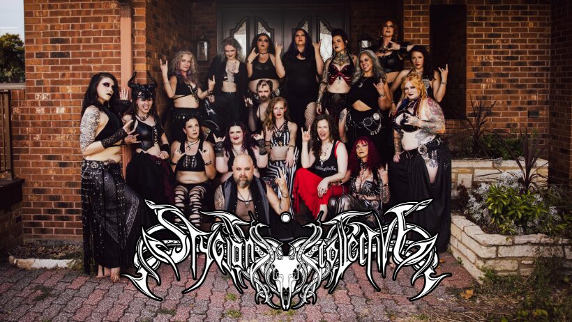 Group of women in gothic clothing posing outdoors in front of a brick building, with a logo at the bottom.